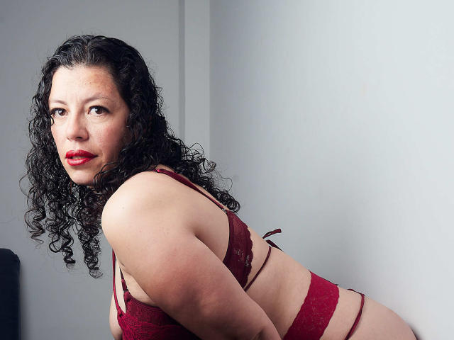ZalyMature, con i capelli neri ricci, rossetto rosso, e reggiseno e slip di pizzo rossi, è china in avanti, mostrando il seno e i glutei. Guarda la fotocamera con un'espressione seria..