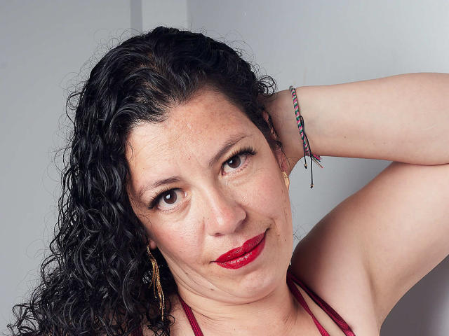 ZalyMature a les cheveux noirs bouclés, du rouge à lèvres, et des yeux marron. Elle porte un haut rouge à encolure américaine et un bracelet coloré, avec son bras levé et un léger sourire..