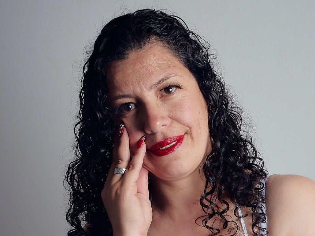 ZalyMature heeft krullend zwart haar, rode lippenstift en een ring aan haar rechterhand. Ze raakt haar wang aan met een lichte glimlach. Ze heeft een lichte huidskleur en donkere ogen..