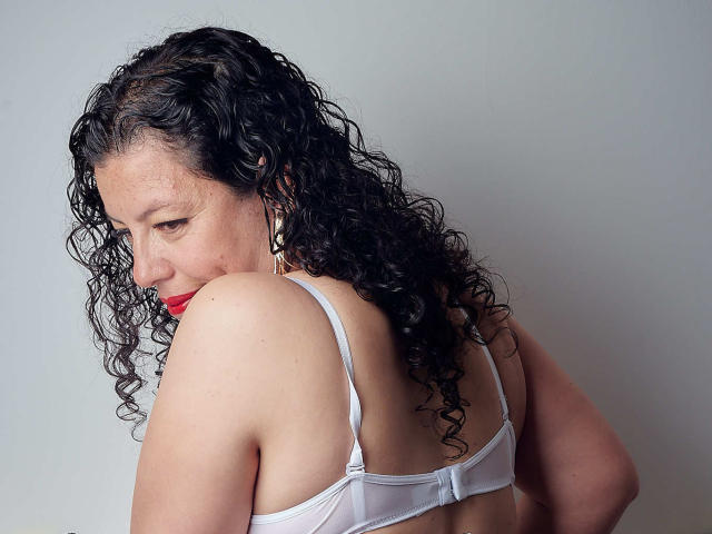 Zaly, con i capelli neri ricci, rossetto rosso e reggiseno bianco, mostra il seno e guarda oltre la spalla, su uno sfondo neutro. Il suo sedere è leggermente visibile..