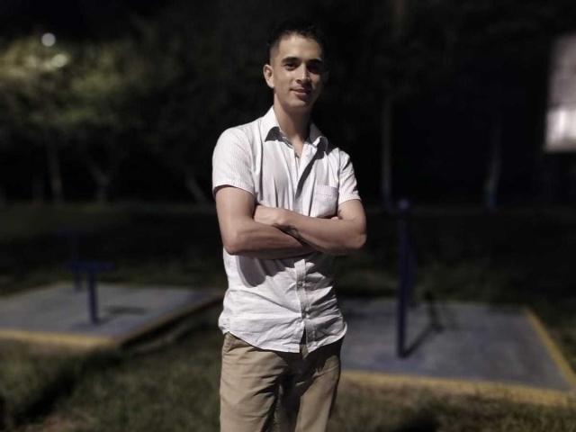 LatinAndres está de braços cruzados, vestindo uma camisa branca de botões, calças bege e uma gola preta. Ele tem uma constituição mediana, cabelo curto e escuro, e um leve sorriso..