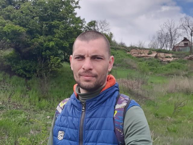 KatyaEgor, schon mit Bart, trägt eine blaue, gepolsterte Weste und ein grünes Hemd, einen Rucksack, steht auf einem grasbewachsenen Feld, lächelt leicht, bewölkter Himmel, ein felsiger Hügel im Hintergrund..