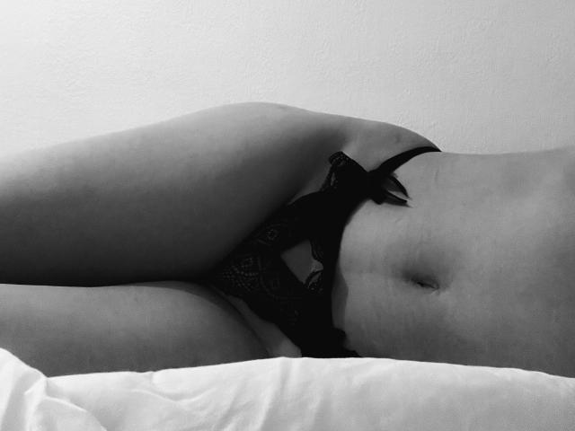 Le mutandine nere in pizzo di Astrxd sono parzialmente abbassate, rivelando il suo torso inferiore, la pelle liscia e chiara, l'addome piatto e la coscia, mentre è sdraiata su un letto bianco in una foto in bianco e nero..