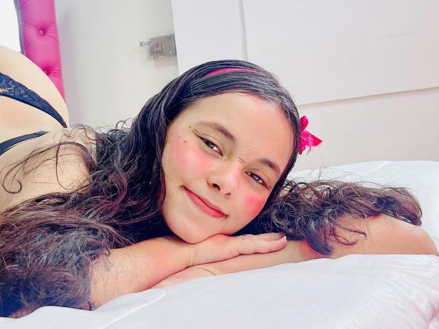 Cloe Ferrara, met lang, krullend zwart haar en een roze hoofdband, ligt op een wit bed, lachend, met roze wangen, en draagt een zwarte kanten bh, terwijl ze haar hoofd op haar hand rust..