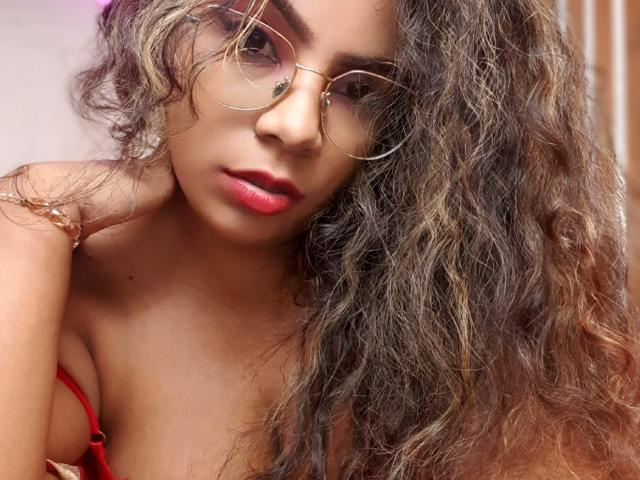 EbonyRose69, con i capelli castano ricci, il rossetto rosso e gli occhiali con montatura dorata, mostra le spalle scoperte e il laccetto rosso del reggiseno. Il seno è leggermente visibile. Sta guardando la fotocamera..