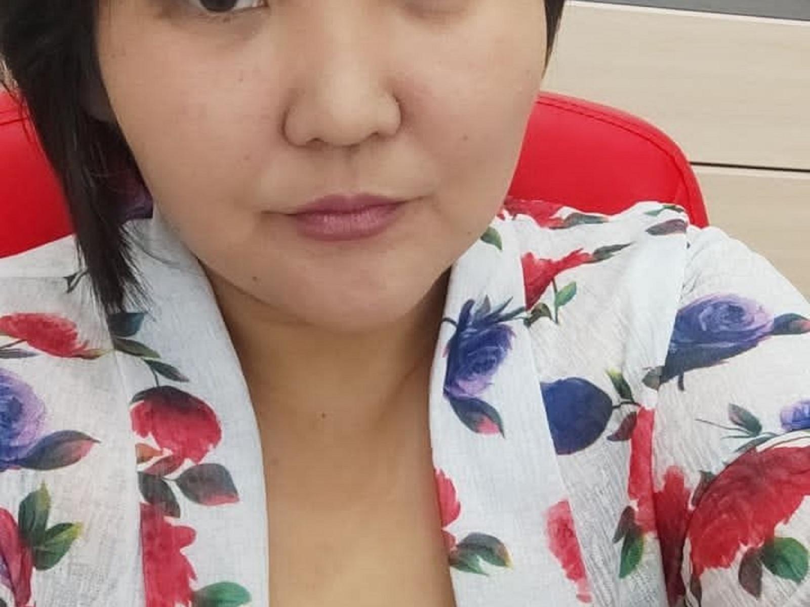 PussyGold with short black hair, light skin, and pink lips, wearing a white floral robe with red, blue, and purple flowers, sitting on a red chair.