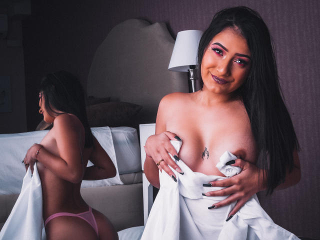 SamyBloom with long black hair, pink eyeliner, and pink thong, covering her tits with a white sheet, smiling at the camera, reflection in mirror showing her ass.