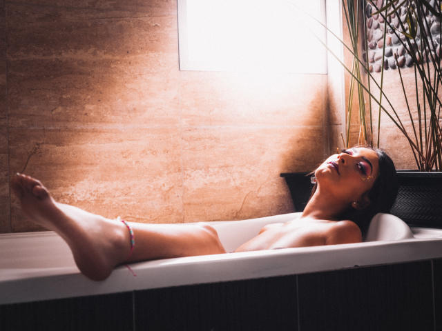SamyBloom está desnuda, con los pechos al descubierto, acostada en una bañera con las piernas levantadas, con maquillaje rosa en los ojos y una pulsera en el tobillo..