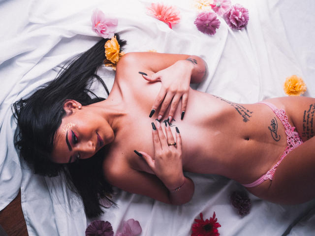 SamyBloom, nue, allongée sur un lit blanc, porte des culottes en dentelle rose, a des ongles noirs, des tatouages sur le côté et le ventre, des fleurs autour d'elle, elle couvre ses petites seins avec ses mains, a les cheveux foncés, et est recouverte d'un drap blanc..