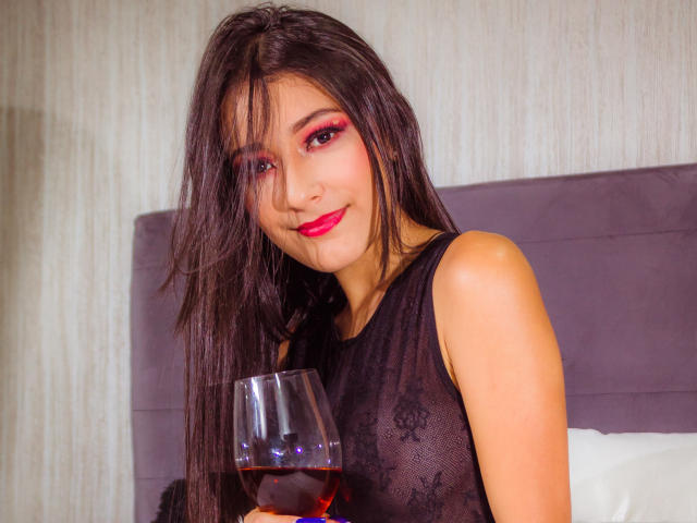 SamyBloom met lang bruin haar, rode oogmake-up, een zwarte kanten top, die een glas rode wijn vasthoudt, en die lichtjes glimlacht. Haar borsten zijn nauwelijks zichtbaar door het kant..