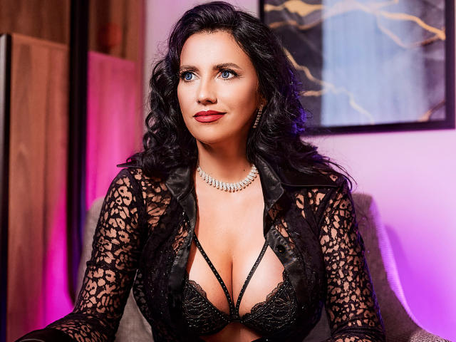 MissFetish with curly black hair, blue eyes, red lipstick, wearing a black lace top with deep cleavage, showing her tits, and a diamond necklace, looking confident.