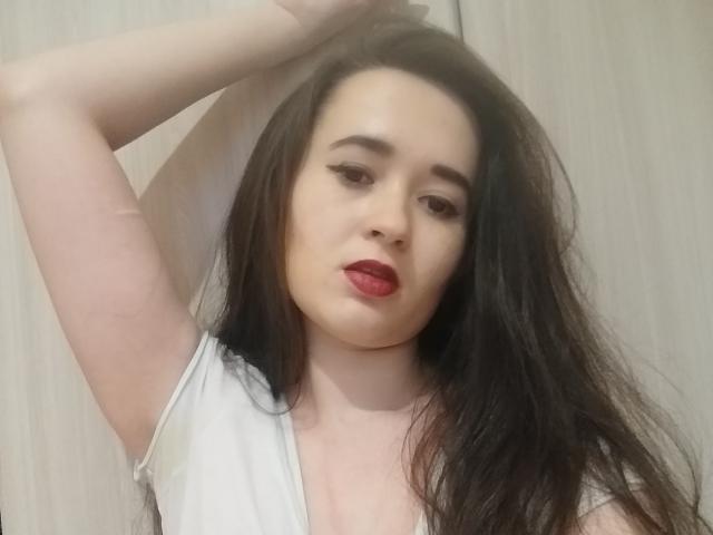 LuxCrow, com cabelo preto longo, batom vermelho, blusa branca, braço levantado, pele clara, olhando para a câmera, delineador discreto, expressão suave, leve lábio franzido, seios de tamanho médio, fundo claro..