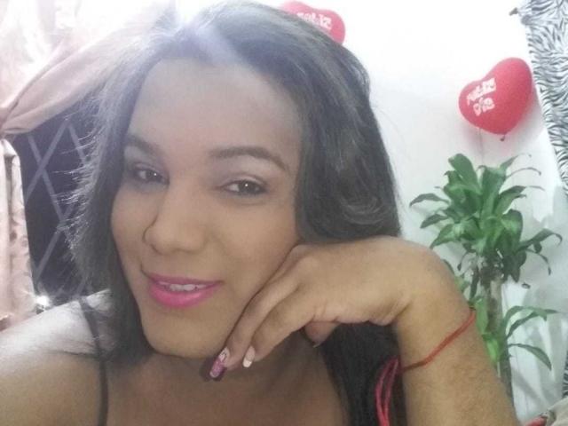 EmilyBigHot mit dunkelbraunen Haaren, pinkem Lippenstift, lächelnd, Kopf auf der Hand, rosa Nägel, rotes Armband, süß, sexy, weiches Licht, leichte Sonnenblendung, rote Herzballons im Hintergrund..
