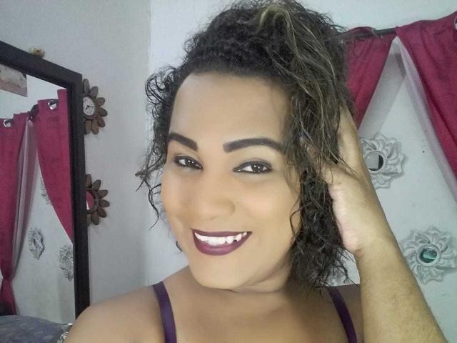 EmilyBigHot hat lockiges, braunes Haar, dunkle Augenbrauen und lila Lippenstift. Sie lächelt mit dunklem, lila Lippenstift, trägt ein lila Oberteil und berührt ihr Haar mit ihrer rechten Hand..