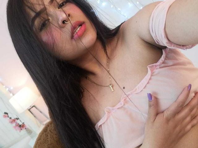 Samantha Velez mit langen, schwarzen Haaren, einem pinken Top mit einem Schulterauschnitt, einem Nasenring, lila Nägeln, einer Hand auf ihrer Brust, leicht geöffnetem Mund, heller Haut und einer Kreuzkette..