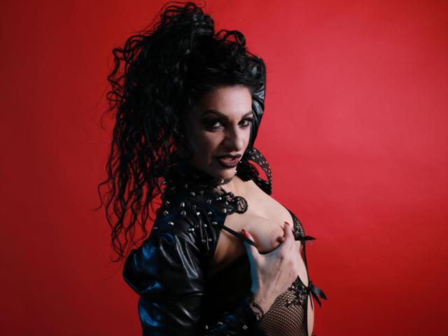 PureWickedGoddess con cabello negro rizado, maquillaje oscuro y una chaqueta de cuero negro, mostrando sus pechos con una mano, sobre un fondo rojo. Sus pezones son visibles..