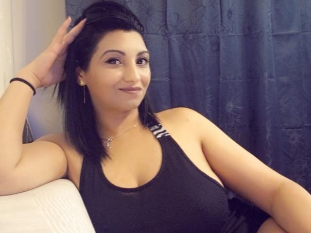 Jessyca, con cabello oscuro, vestida con una camiseta deportiva negra, mostrando sus pechos, sentada, sonriendo, con la mano en el pelo, cortina azul, cama blanca, pulsera negra, aretes dorados..