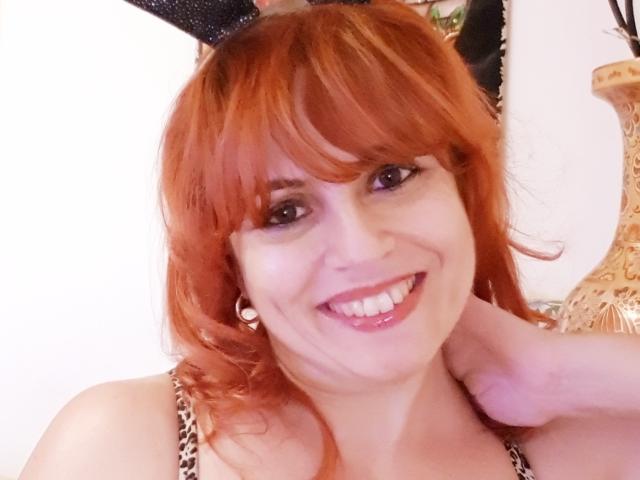 HotHarissa: Capelli rossi, orecchie da coniglio nere e scintillanti, sorridente, con i denti in mostra, indossa una maglietta a stampa leopardo, una mano al collo, indossa orecchini d'argento ed è molto carina..