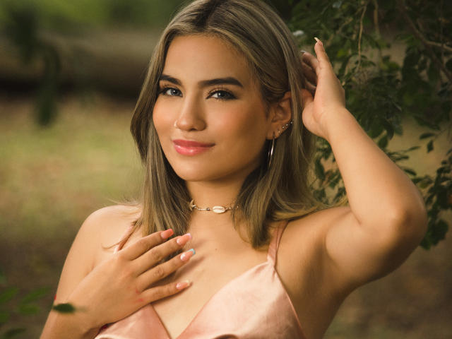 Celeste Bennett, com cabelo loiro, vestido rosa e colar de ouro, tocando o peito com a mão, sorrindo, olhos verdes, maquiagem discreta, fundo de floresta, mão no cabelo, brincos..