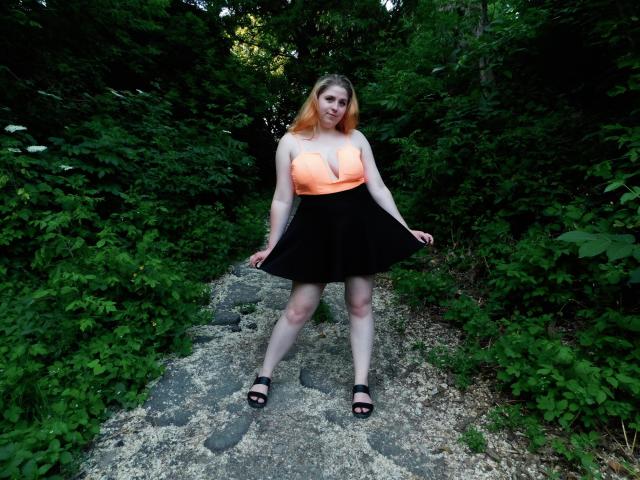 JessikaDaniels has pale skin, orange hair, and is wearing a neon orange top with black skirt, showing her tits, black sandals, standing on a rocky path in forest.