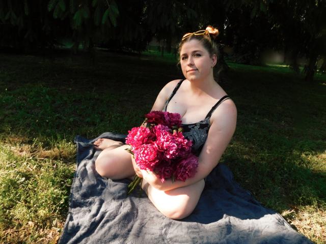 JessikaDaniels, sitting on a dark blanket, holding a bouquet of deep pink flowers, wearing a black lace dress, with her tits and ass slightly visible, in a grassy area.