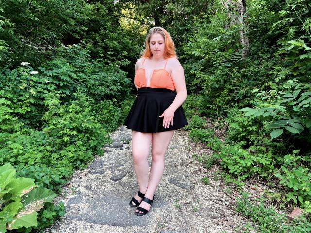 JessikaDaniels has red hair, orange strapless top, black skirt, and black sandals. She stands on a stone path, surrounded by green foliage, looking down with a slight smile.