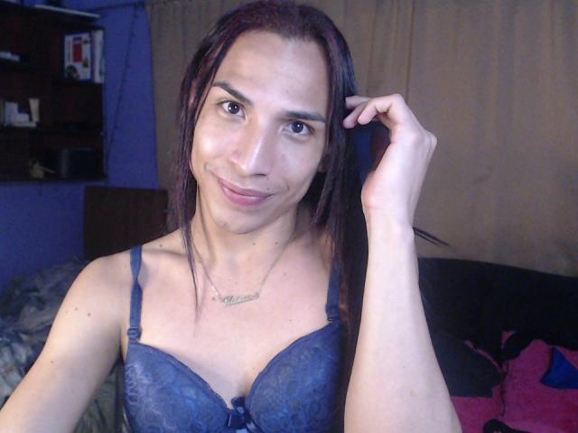 Brunettehot77 with long dark hair, wearing a blue lace bra, smiling at the camera, hand in hair, necklace with cursive writing, in a dimly lit room.