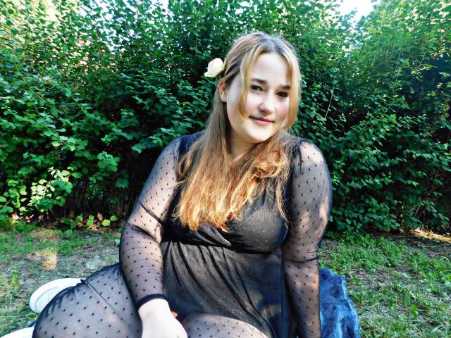 DaniellaGraceZZ with light brown hair, a small white flower in her hair, sitting on grass, wearing a black, sheer, polka-dot dress, smiling, showing her ass slightly.