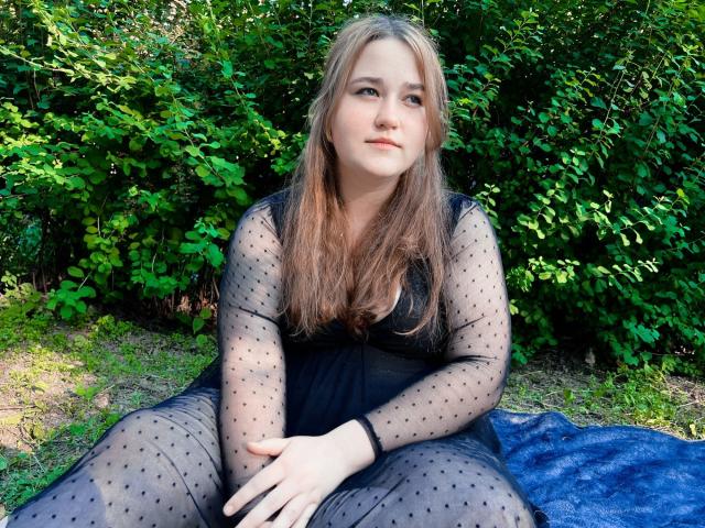 DaniellaGraceZZ, with long blonde hair, sits on a blue blanket, wearing a sheer black dress with polka dots, hands on lap, green bushes in the background.