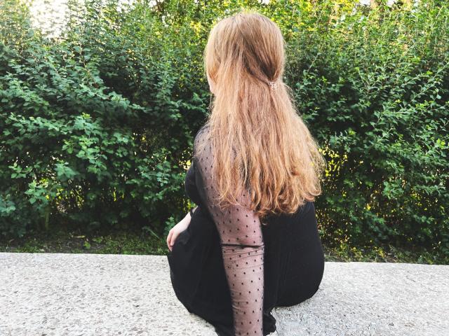 DaniellaGraceZZ, sitting on a concrete surface, back to the camera, with long, wavy, blonde hair, wearing a black sheer polka-dot top, and black dress, in a green bush setting.