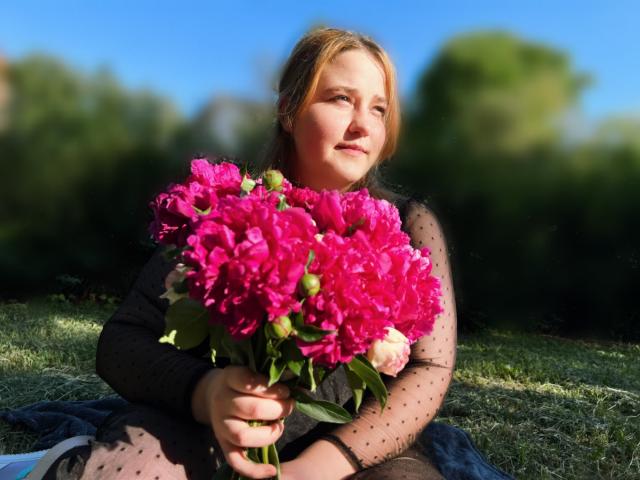 DaniellaGraceZZ with pink peony bouquet, wearing black sheer dress, sitting on grass, holding flowers, blue sky, green trees, smiling, sunlight, blonde hair, cute, soft light.