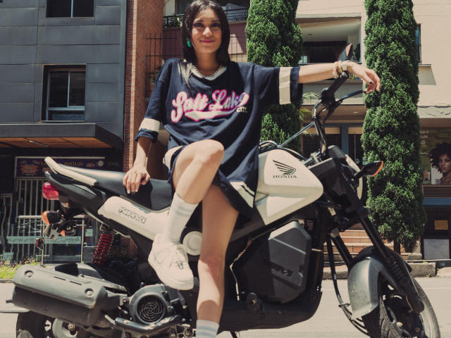 MeliMuller sits on a white Yamaha motorcycle, wearing a navy shirt with pink "Jet" text, white socks, and sneakers, with one leg up, in a sunny urban setting.
