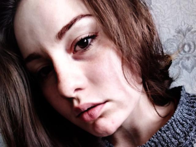 KrissySky's face in a tight shot, brown wavy hair, slightly parted lips, brown eyes with subtle makeup, wearing a gray knit sweater, looking tired and pensive.