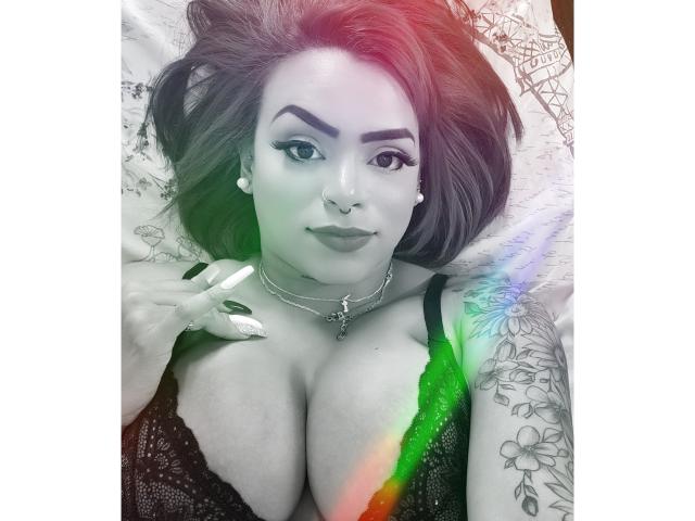AngelFallon lying on a bed, green light on her tits, wearing black lace bra, nose ring, and pearl earrings, with tattooed arm and rainbow light.