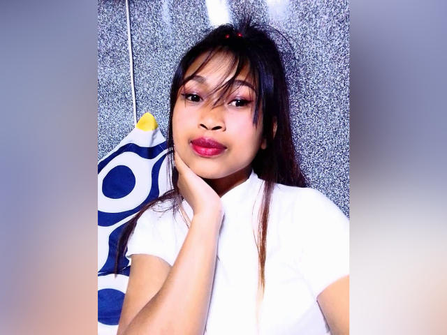 AnnahMaru, avec ses cheveux foncés, son rouge à lèvres rose et sa chemise blanche, touche sa joue, portant un vêtement bleu et blanc à motifs suspendu derrière elle. Ses seins sont petits et mignons..
