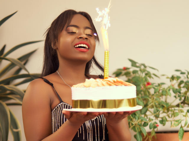 LindaBird with dark skin, holding a birthday cake with a lit candle, wearing a black and white dress, smiling with red lipstick, and medium-sized tits, in a photo.