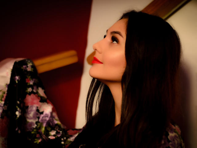 AshlyChloe with long black hair, red lipstick, and a floral dress, looking up with a slight smile, soft light highlighting her face, subtle nose piercing.