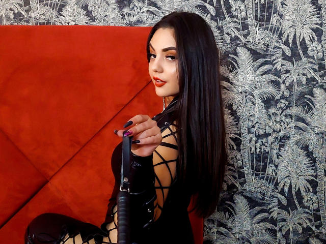 AshlyChloe, con cabello negro largo, vestida con un traje de látex negro, sosteniendo una paleta de cuero negro, sentada en un sofá rojo, mirando hacia atrás con una ligera sonrisa..