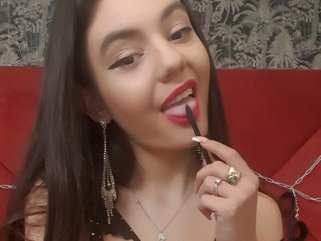 AshlyChloe, avec du rouge à lèvres rouge, du crayon noir pour les yeux et de longs cheveux noirs, léchant un applicateur de crayon noir, portant des boucles d'oreilles pendantes et une bague, souriant. Les seins sont partiellement visibles, fond rouge..
