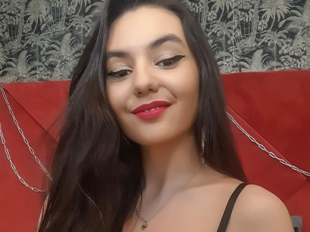 AshlyChloe, con lunghi capelli scuri, rossetto rosso, eyeliner nero, sorridente, indossa un top nero con spalline e una collana. Ha il seno piccolo e un viso carino..