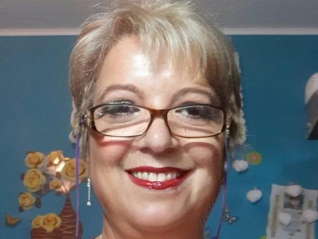 QueenPammy with short blonde hair, glasses, red lipstick, smiling, wearing purple earrings, blue wall, yellow floral decor, and a white ceiling. Her tits are not visible.