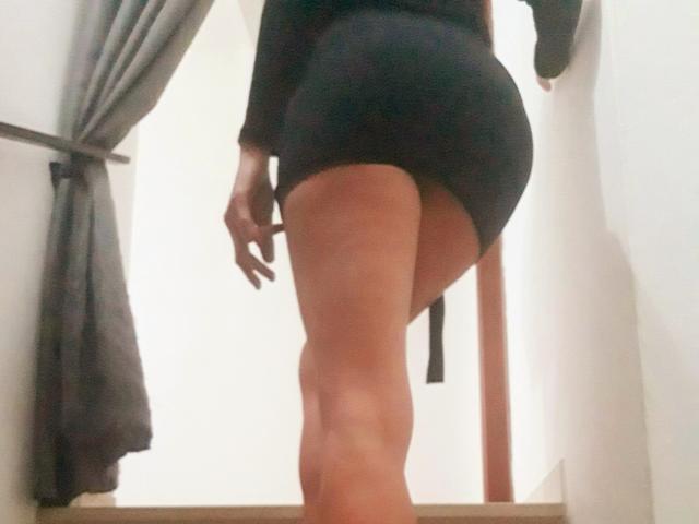 Les fesses et les cuisses d'Aiyana, vêtue d'une robe noire moulante, debout sur un plancher en bois, face à l'arrière-plan, avec des rideaux gris. Sa main est partiellement visible..