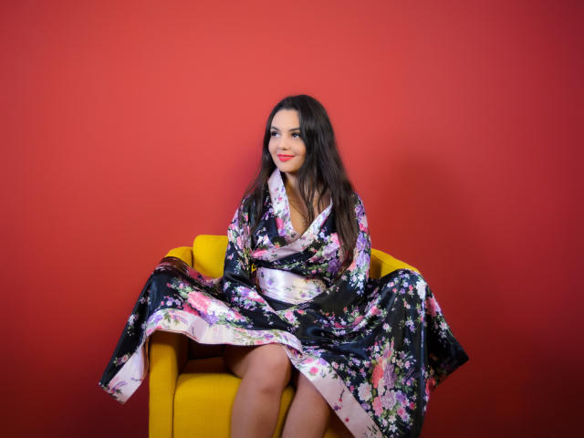 AshlyChloe, with long dark hair and red lipstick, sits on a yellow chair, wearing a black floral kimono, exposing her bare ass, against a red wall.