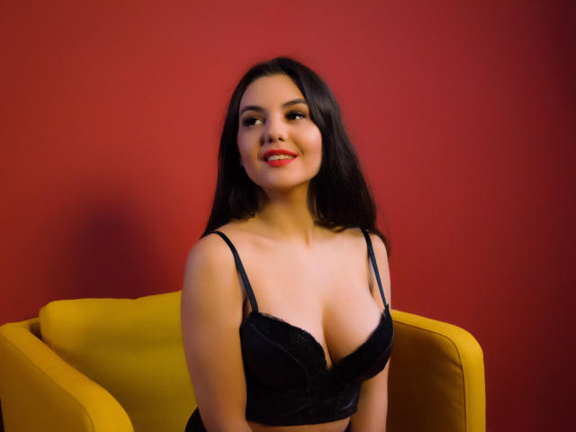 AshlyChloe with long dark hair, smiling, wearing a black lace bra, showcasing her tits, sitting in a yellow chair against a red wall, looking cute and sexy.