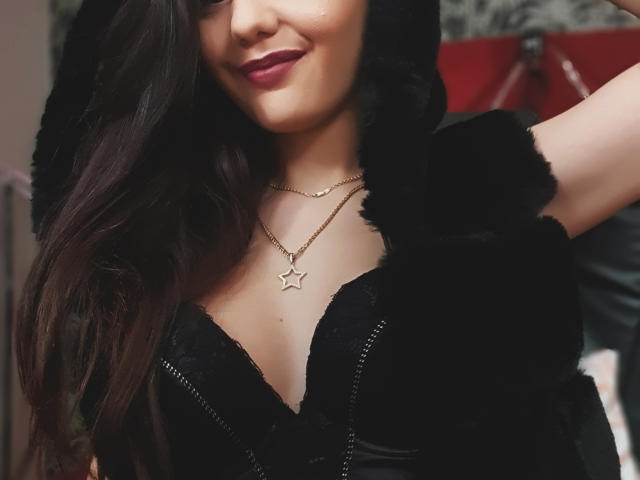 AshlyChloe met donkerbruin golvend haar, een zwarte hoodie met imitatiebontvoering en een sterrenketting, lachend, waarbij haar borsten zichtbaar zijn in een zwarte kanten BH. Ze heeft roze lippenstift op..