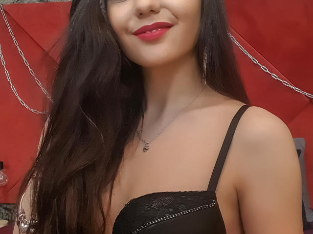 AshlyChloe has long black hair, red lipstick, and wears a black lace bra. She's smiling with her tits visible and a silver necklace. Background is red with chains.