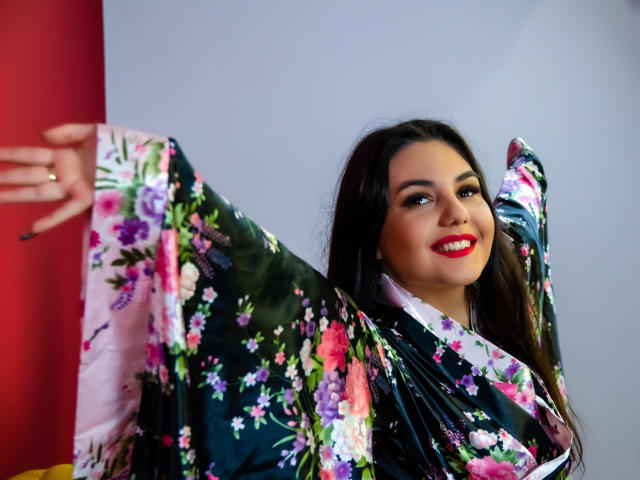 AshlyChloe in a floral kimono, arms out, smiling with red lipstick, dark hair, green eyes, wearing a ring, black kimono with pink, purple, and white flowers.