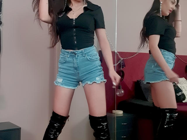 AshlyChloe with long dark hair, black tight shirt, blue distressed shorts, black thigh-high boots, and a chain necklace, standing in front of a mirror reflecting her ass.