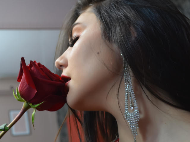 AshlyChloe, avec ses longs cheveux noirs, une rose rouge dans la bouche, portant des boucles d'oreilles scintillantes, a une peau pâle et une petite tache de naissance près du nez. Elle est séduisante et regarde sur le côté..