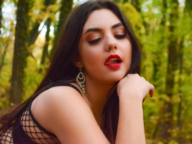 AshlyChloe met rode lippen, donkere make-up en lang zwart haar, draagt een nethemd en gouden oorbellen, leunt haar kin op haar hand en ziet er sexy uit in een bos..